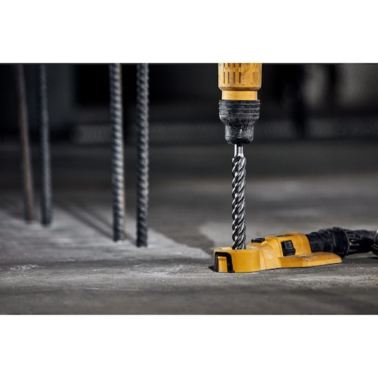 A close-up view of a DEWALT drill with a spiral drill bit attached, actively drilling into a concrete floor. The tool is yellow and black, and nearby steel rebar rods are visible in the background.