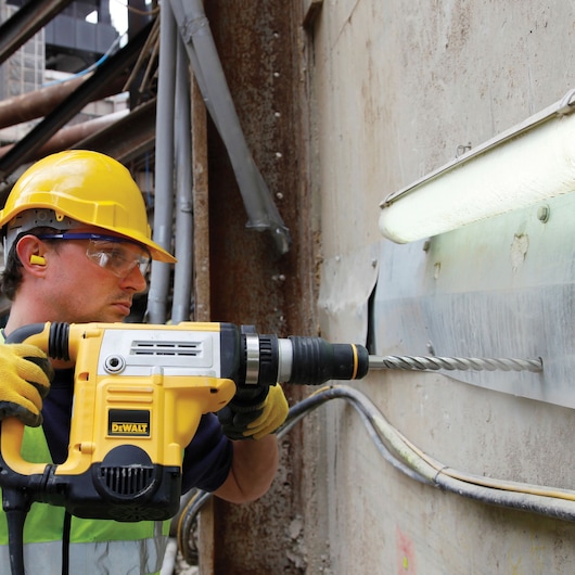 A worker wearing a yellow hard hat, ear protection, and gloves uses a DEWALT rotary hammer drill to bore into a concrete wall at an industrial site.