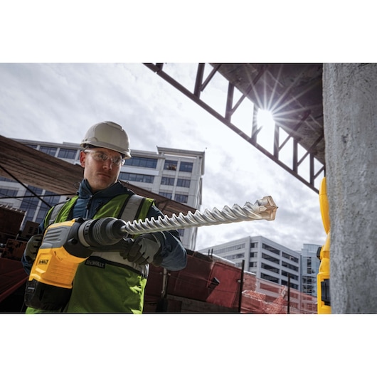 A person wearing a construction helmet and high-visibility vest is using a DEWALT power tool with a long, spiral drill bit at a construction site, with modern buildings in the background.