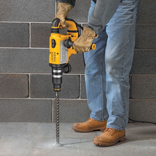 A person wearing work gloves, jeans, and boots is using a yellow DEWALT hammer drill to drill into a concrete floor. The drill has a long, spiral bit and is being operated near a grey brick wall.
