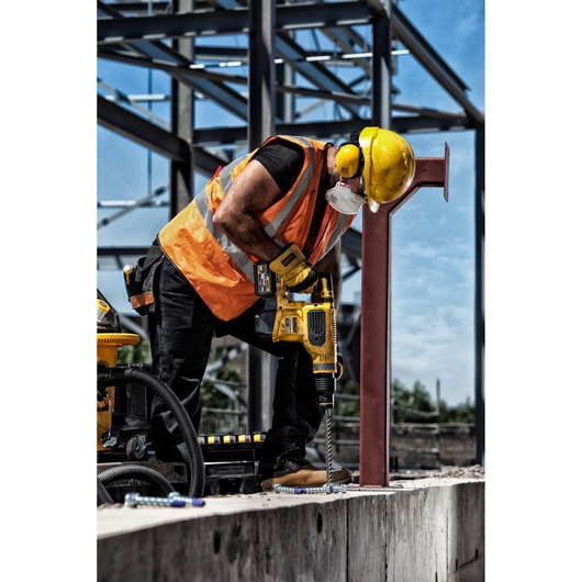 A construction worker wearing a yellow hard hat, orange safety vest, and face mask is using a Dewalt cordless rotary hammer drill to drill into a concrete surface at a construction site. Steel frameworks are visible in the background.