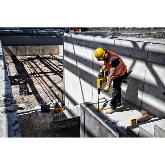 A construction worker wearing a yellow helmet and orange safety vest is using a DEWALT power drill to drill into a concrete surface at a construction site. Various tools and equipment are visible on the ground nearby.