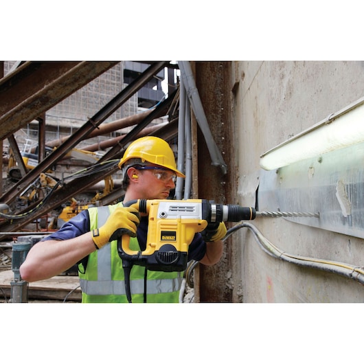 A construction worker wearing a yellow hard hat, safety vest, ear protection, and gloves is using a DEWALT electric drill to drill into a concrete wall at a construction site. The scene includes exposed pipes and structural beams.