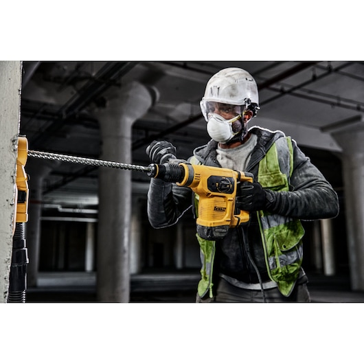 A construction worker wearing a helmet and safety vest is using a yellow DEWALT rotary hammer drill to drill into a concrete wall in an industrial setting.