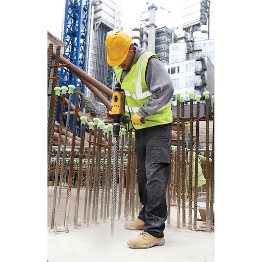 A construction worker wearing a yellow hard hat and high-visibility vest is using a DEWALT power drill at a building site. Reinforcing steel bars and urban buildings are visible in the background.