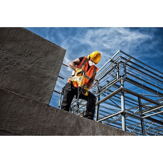 A construction worker wearing a yellow hard hat, orange safety vest, and face mask operating a yellow DEWALT power tool on a concrete slab at an industrial building site with steel framework visible in the background.