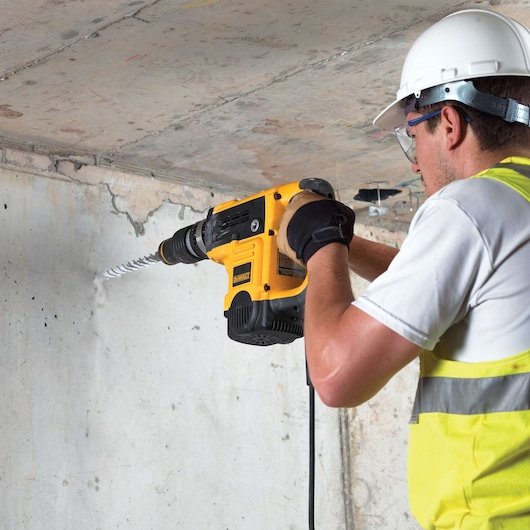 A construction worker wearing a white hard hat, safety glasses, gloves, and a yellow vest is using a DEWALT electric drill with a long bit to bore into a concrete wall.
