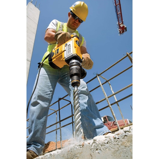 A construction worker wearing a hard hat and gloves uses a DEWALT power drill with a large masonry bit to drill into concrete at a construction site. Rebar and a crane are visible in the background.