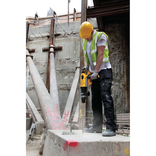 A construction worker wearing a yellow hard hat and reflective safety vest is using a DEWALT rotary hammer drill on a concrete block at a construction site.