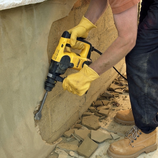 A person wearing yellow gloves and tan work boots uses a yellow DEWALT power tool to chip away plaster on a wall, with debris falling to the ground.