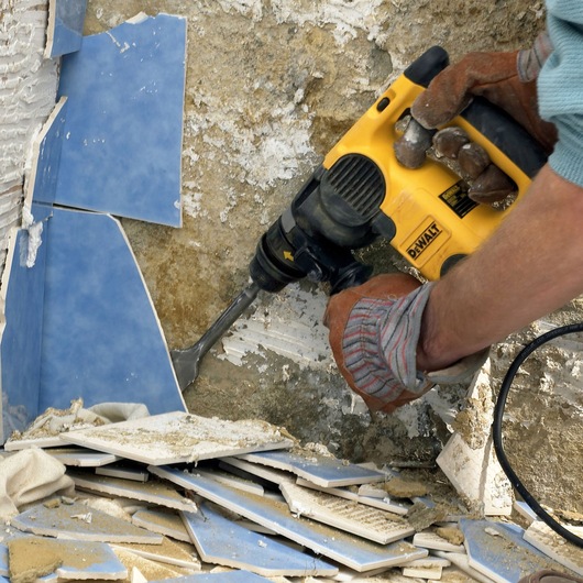 A person wearing work gloves uses a yellow DEWALT power tool to remove blue tiles from a wall. Broken tiles are scattered on the ground.