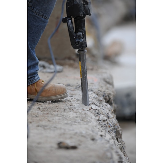 Worker uses a DEWALT chisel tool to break concrete on a construction site, wearing jeans and safety boots.