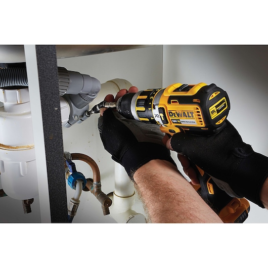 A person wearing black gloves uses a DEWALT cordless drill to fasten a pipe connection under a sink.