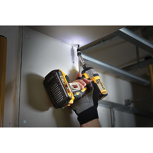 A person wearing work gloves uses a DEWALT cordless power drill to drive a screw into a ceiling corner, with a built-in LED light illuminating the work area.