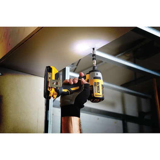 A person wearing a glove uses a DEWALT cordless power drill to drive a screw into an overhead surface. The drill has an LED light illuminating the work area.