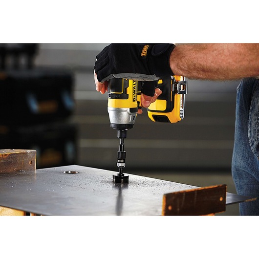 A person using a yellow and black DEWALT power drill with a hole saw attachment to drill through a metal sheet, wearing protective gloves.