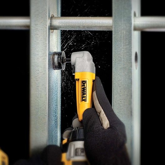 A person wearing work gloves uses a yellow DEWALT right angle attachment tool to drill into a metal beam, with sparks visible during the drilling process.