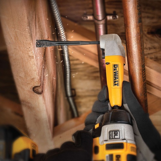 A gloved hand using a DEWALT drill with a drill bit attachment to work on a wooden beam in a construction setting. Electrical and copper pipes are visible nearby.
