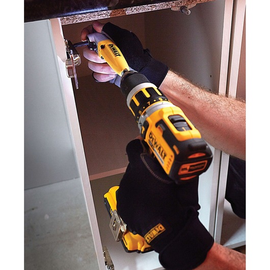 A person wearing black gloves is using a yellow and black DEWALT cordless drill to install or tighten a hinge inside a cabinet.