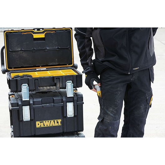 A person wearing black work clothing is standing next to a stack of DEWALT toolboxes, with the top toolbox open to reveal yellow compartments for organizing tools or accessories. The person is holding a DEWALT item in their hand.