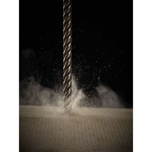 A close-up view of a DEWALT spiral drill bit penetrating a concrete surface, creating dust and debris against a dark background.