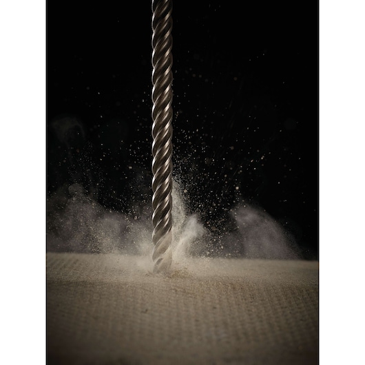 Close-up of a metallic drill bit penetrating a surface, with dust and particles being scattered, against a dark background.
