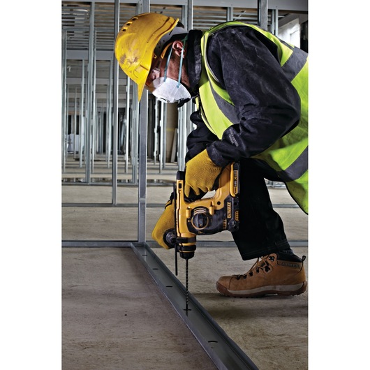 A construction worker wearing a yellow hard hat, high-visibility vest, gloves, and a face mask is kneeling while using a DEWALT power drill (model DT8926) to drill into a metal frame on a construction site.