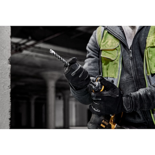 A person wearing work gloves and a green safety vest is holding a DEWALT power drill with a spiral drill bit attached, preparing for use in an industrial or construction setting.