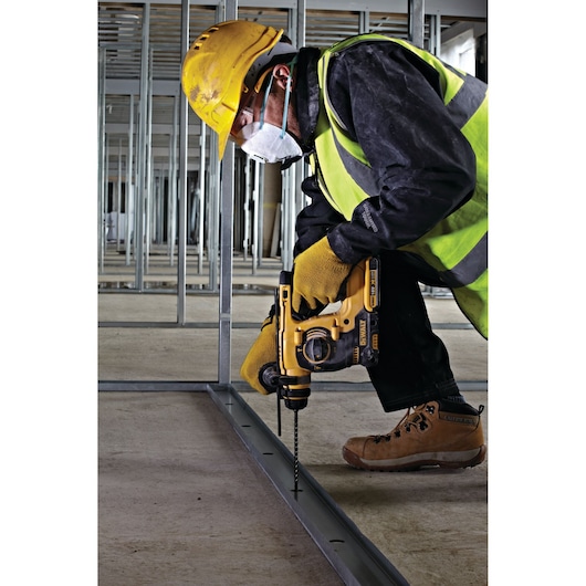 Person wearing a yellow hard hat, high-visibility safety vest, gloves, and protective mask using a DEWALT power drill to drill into a metal frame on a construction site.