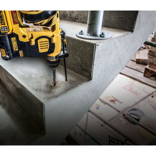 A close-up of a DEWALT power drill drilling into a concrete staircase indoors, with some metal construction elements visible.