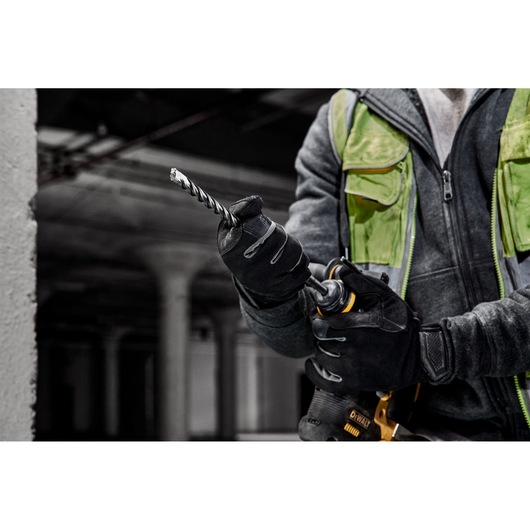 A person wearing black gloves and a green utility vest is holding a DEWALT power drill fitted with a spiral drill bit, in an industrial indoor environment.