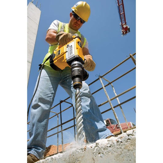 A construction worker wearing a yellow safety helmet and gloves is using a DEWALT rotary hammer drill with a large auger bit to drill into concrete at a building site. Steel rebar, a crane, and construction materials are visible in the background.