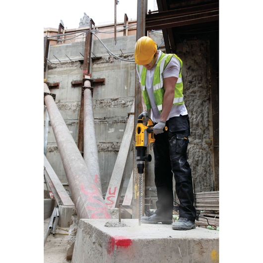 A construction worker wearing a yellow hard hat and a reflective vest is using a DEWALT rotary hammer drill to drill into a concrete block at a construction site.