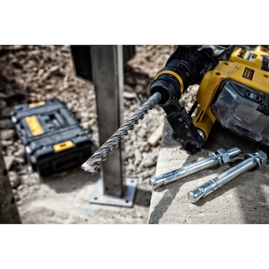 A close-up of a yellow DEWALT rotary hammer drill with a masonry drill bit attached, lying on a concrete surface next to three metal anchor bolts. In the background, a black DEWALT tool case is visible on a construction site.