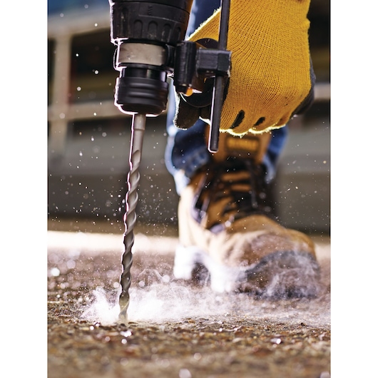 Close-up of a person wearing a yellow work glove using a power drill with a masonry bit to drill into a concrete surface, with dust and debris scattering.