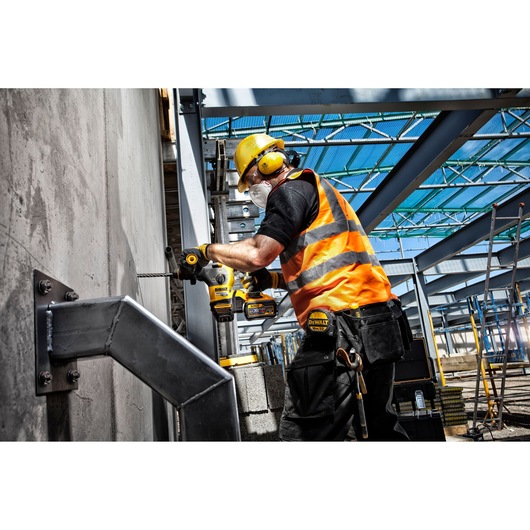 A construction worker wearing safety gear, including a yellow hard hat, ear protection, safety glasses, mask, and an orange reflective vest, is using a DEWALT cordless drill to drill into a concrete wall at a construction site with steel beams.