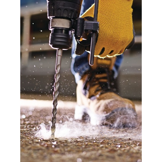 Close-up of a person wearing yellow work gloves and work boots using a drill with a masonry bit to drill into a concrete surface, with dust particles visible around the area.