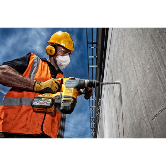 A construction worker wearing a yellow hard hat, orange safety vest, and yellow gloves is using a DEWALT XR cordless rotary hammer drill to drill into a concrete wall outdoors under a blue sky.