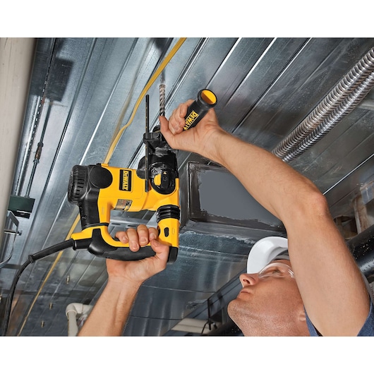 A person using a DEWALT rotary hammer drill to drill into a metal ceiling. The drill is yellow and black, and the person is holding the tool with both hands while working overhead.