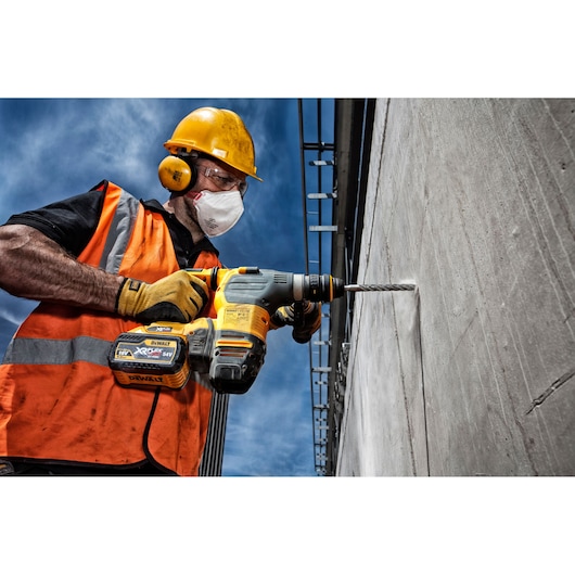 A construction worker wearing a yellow hard hat, orange safety vest, and gloves is using a DEWALT cordless rotary hammer drill to bore into a concrete wall outdoors under a blue sky.