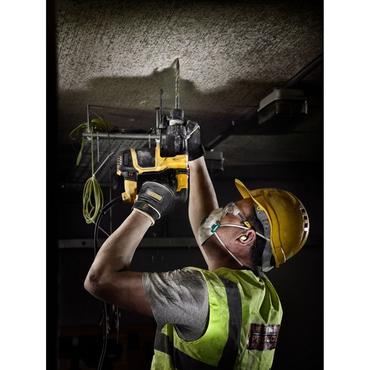A construction worker wearing safety gear uses a DEWALT electric drill to bore into a ceiling. The worker is equipped with a hard hat, gloves, high-visibility vest, dust mask, and ear protection.