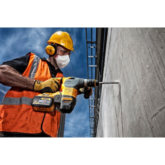 A construction worker wearing a yellow hard hat, orange safety vest, and gloves is operating a DEWALT cordless rotary hammer drill to drill into a concrete wall outdoors.