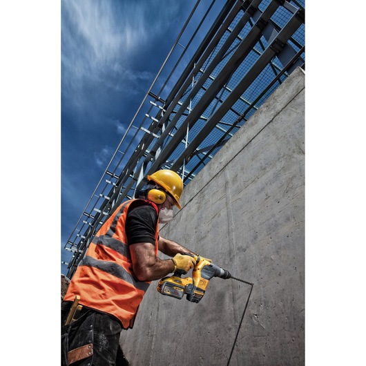A construction worker wearing an orange safety vest, yellow hard hat, ear protection, gloves, and a mask is using a yellow DEWALT cordless rotary hammer drill to drill into a concrete wall outdoors, with a blue sky and metal structure in the background.