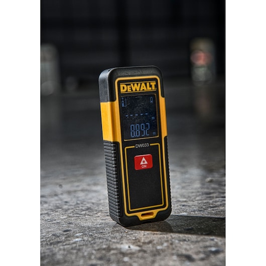 A DEWALT DW033 laser distance measurer standing upright on a concrete surface, with a digital display showing measurement readings and a prominent red ON button.