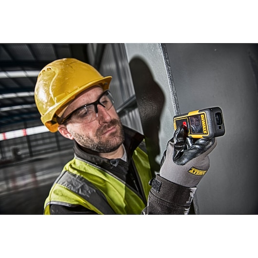 A person wearing a yellow hard hat, high-visibility vest, and gloves is using a DEWALT DW033 laser distance measurer against a metal surface in an industrial setting.