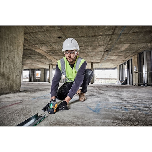 A person wearing a safety helmet, high-visibility vest, and gloves is kneeling on the floor of a large construction site