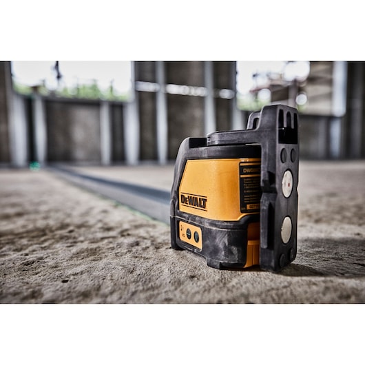 A DEWALT DW088CG green cross line laser level device placed on a concrete floor, with a visible green laser beam projecting across an unfinished construction site.