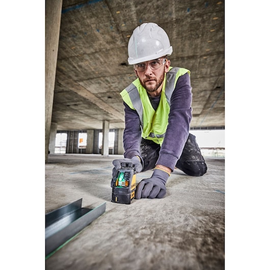A construction worker wearing a hard hat and safety vest is using a DEWALT DW088CG green cross line laser level on a concrete floor at a building site.