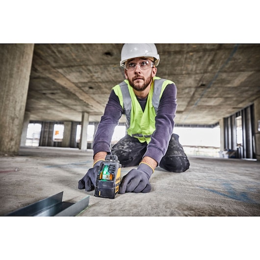 A construction worker wearing a hard hat and safety vest is kneeling on a concrete floor, using a DEWALT DW088CG green cross line laser tool. The tool is placed on the ground to assist in alignment and leveling during building construction.