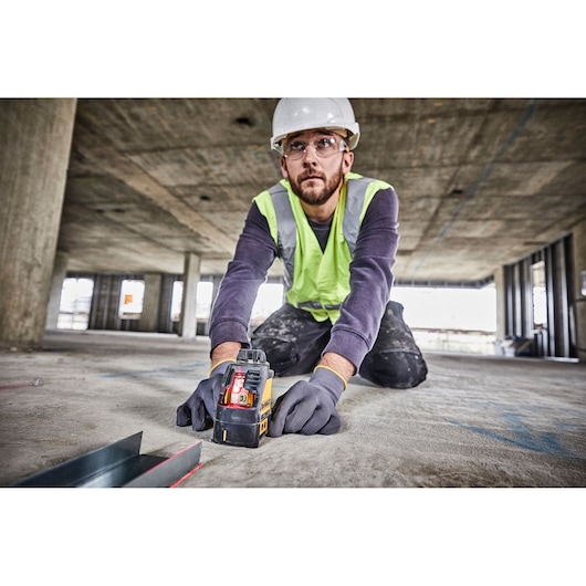 A person wearing a hard hat, safety vest, and gloves is kneeling on the floor of a construction site, using a DEWALT DW088K laser level. The device is projecting a visible red laser line for alignment. The surroundings show an unfinished building with concrete columns and open windows.